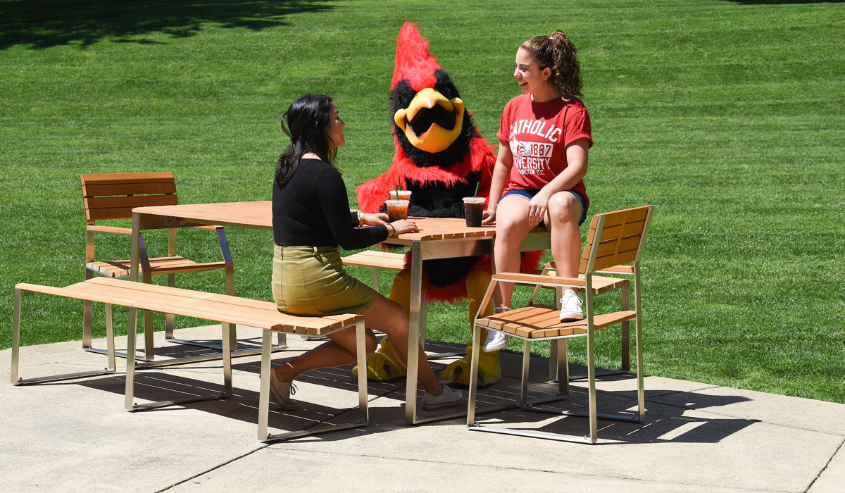 Female students sitting outside and talking to Red the mascot